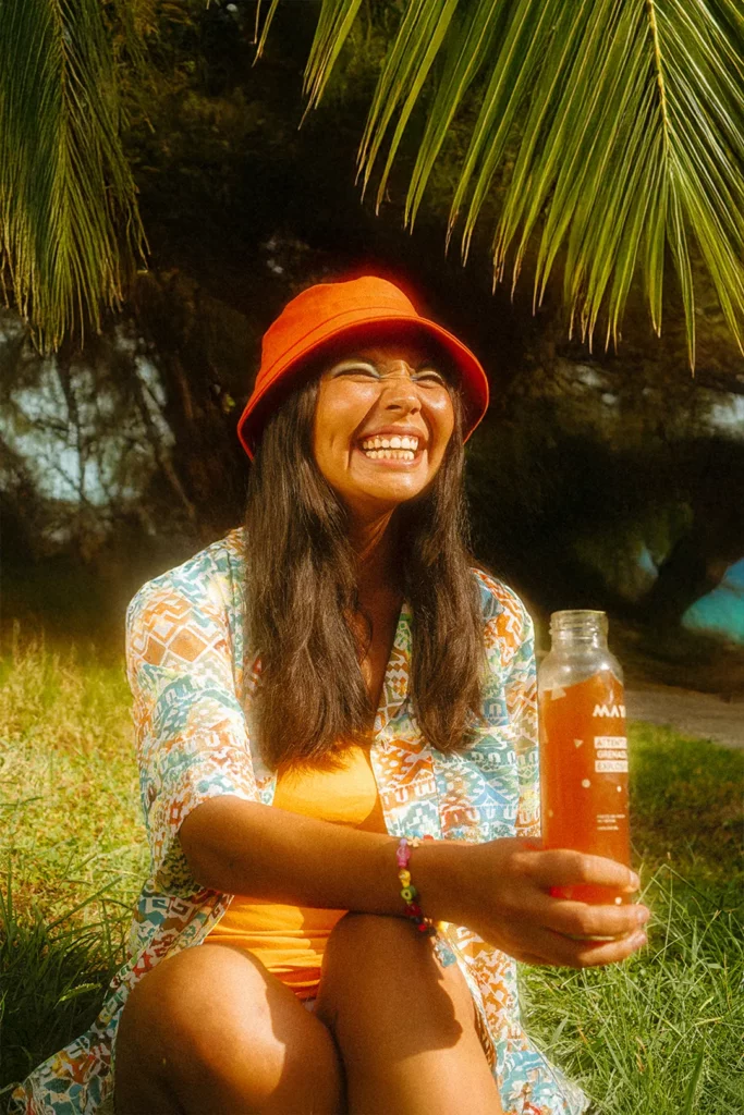 Femme riant assise dans l’herbe entre les palmiers, boisson Maya à la main, lumière douce d’été
