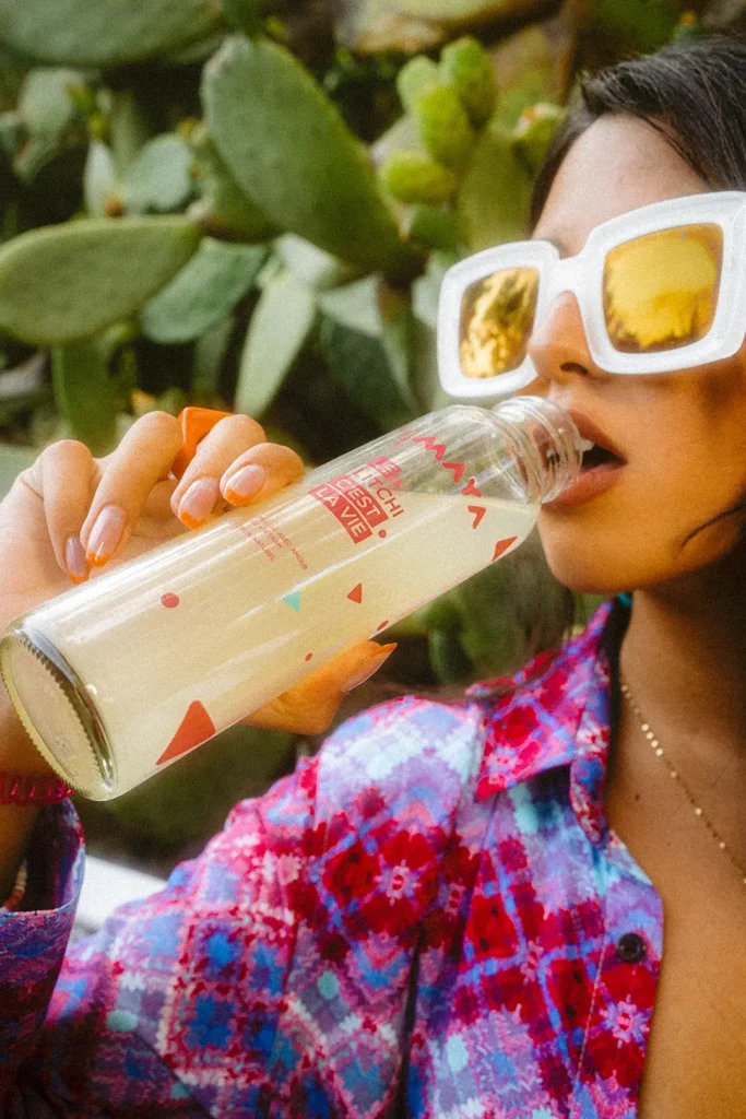 Femme de 3/4 face portant lunettes vintage, sirote une boisson Maya au litchi, cactus en arrière-plan