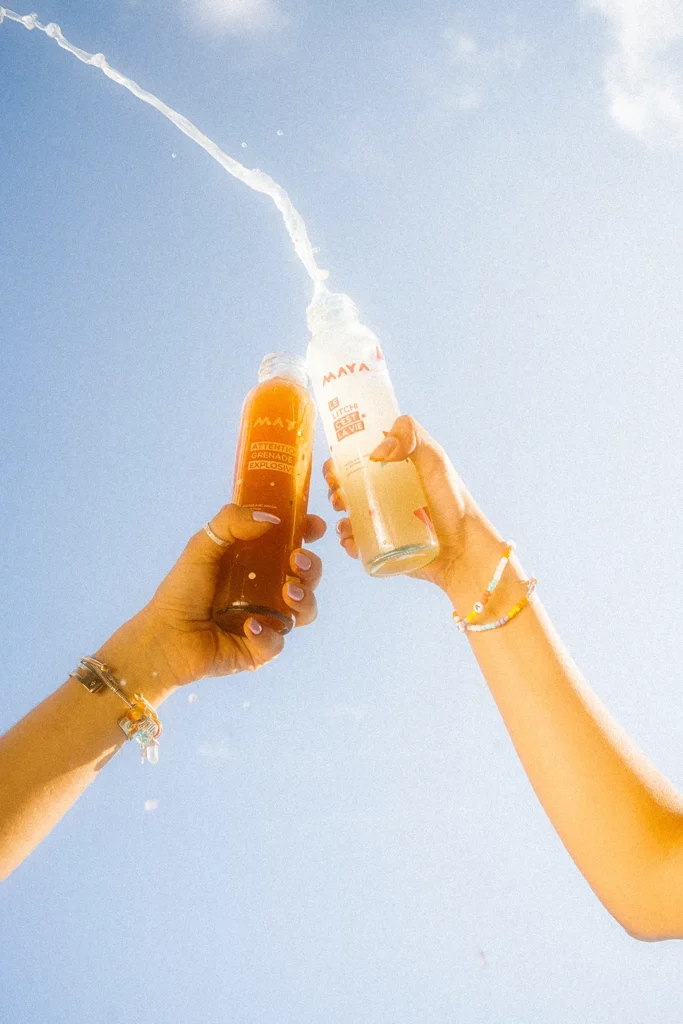 Deux mains trinquent avec des bouteilles Maya, éclaboussures de jus sous ciel bleu, énergie estivale
